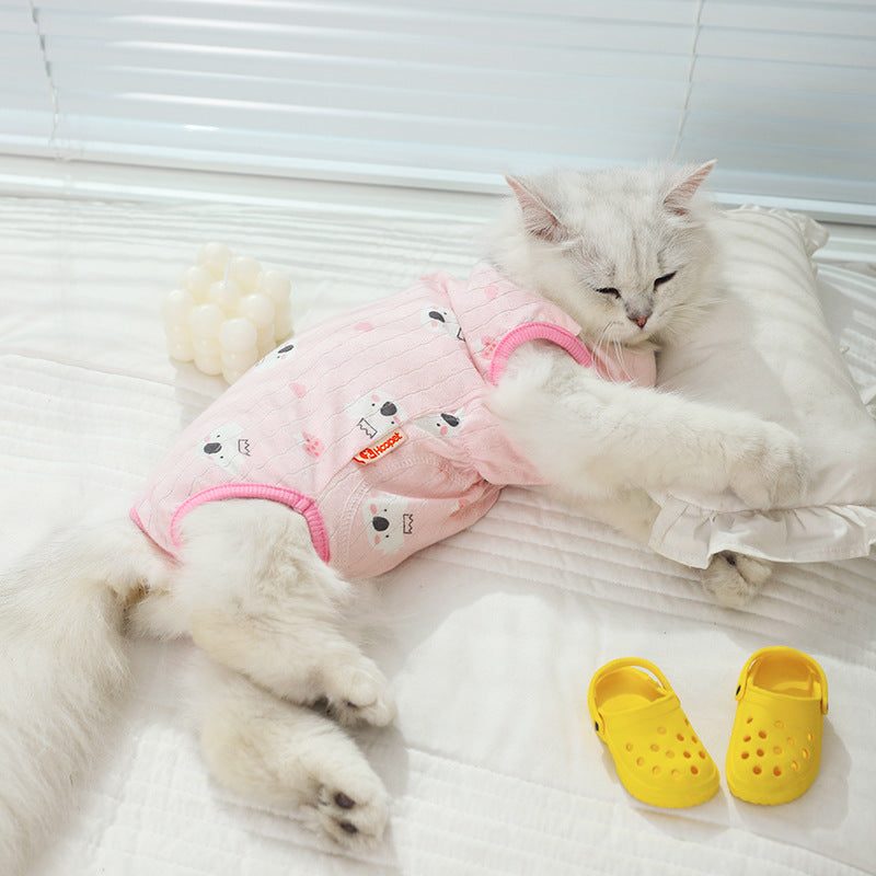 Cat wearing pink anti-shedding clothes, lying on a bed with a pillow and yellow shoes nearby.